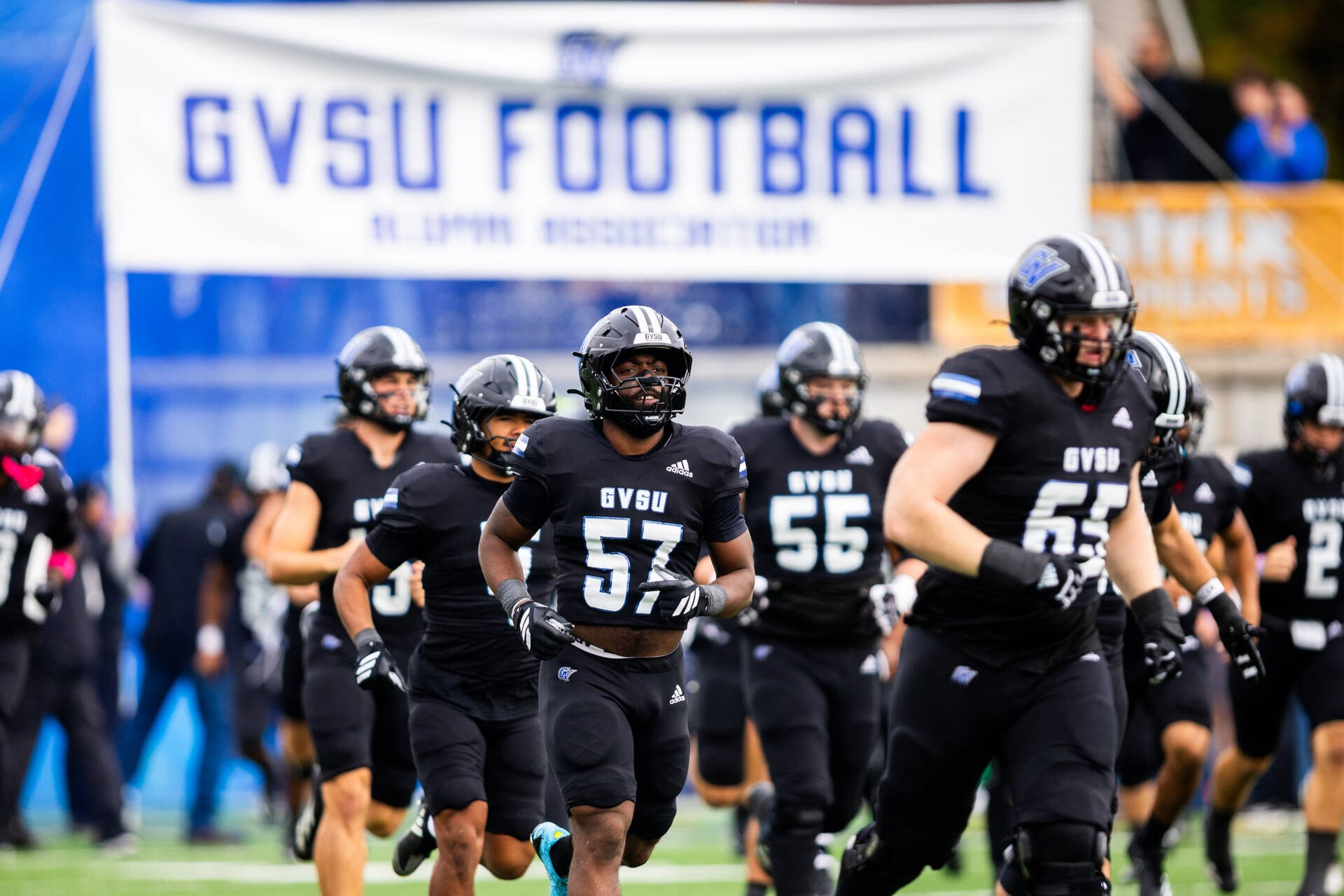 Laker football team runs under GVSU Football banner wearing all black with black helmets
